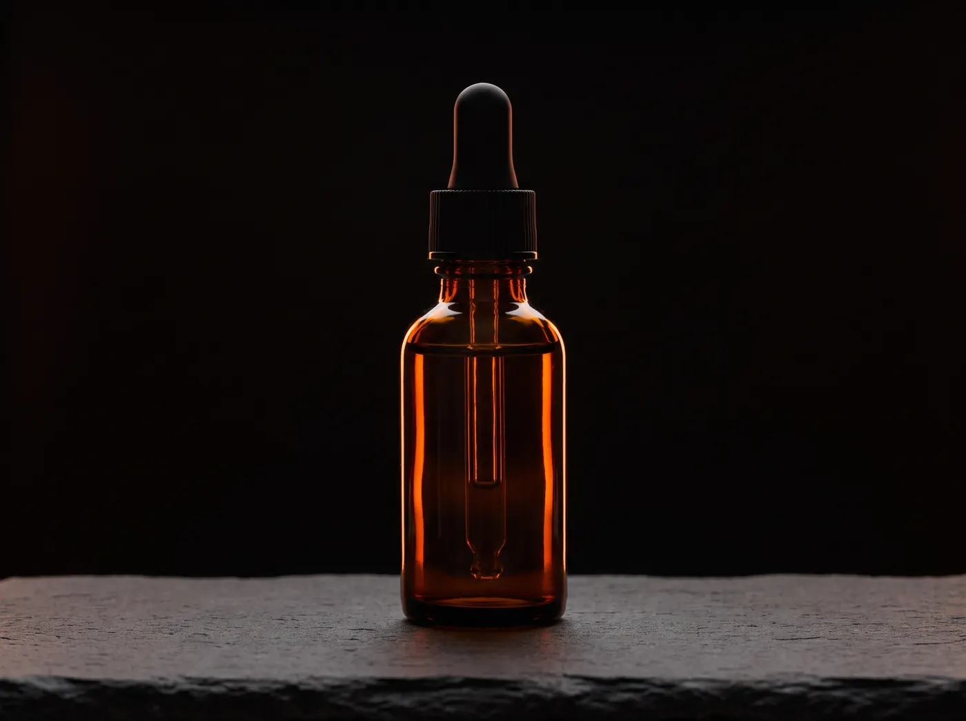 An unlabeled amber dropper bottle on dark slate under warm light